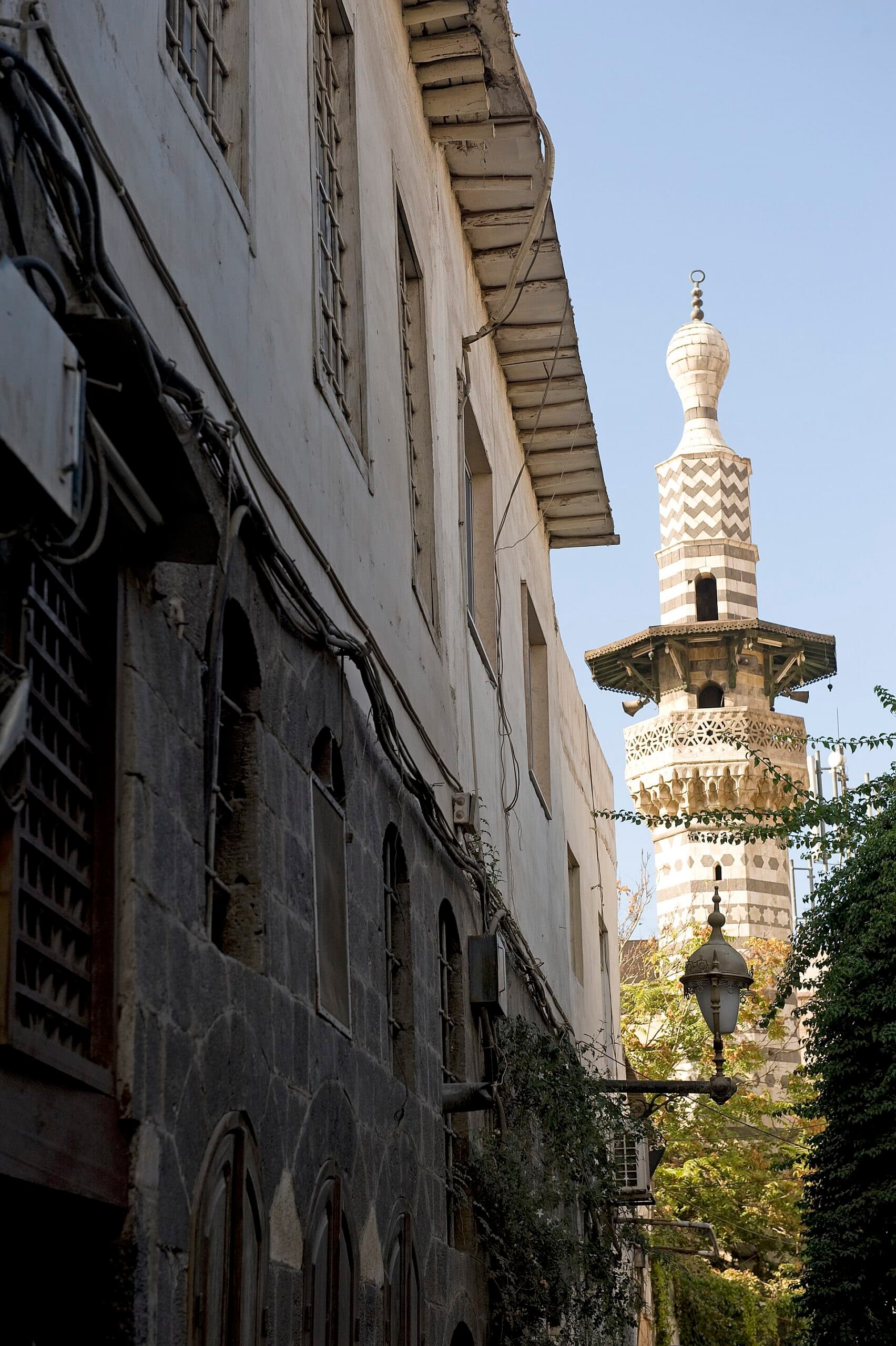 Şam'da Kaymeriyye'nin dar bir sokağının ucunda yükselen Safercelani Camii'nin siyah beyaz taş desenli sekizgen minaresi; solda ahşap saçaklı, kafesli pencereli eski bir ev