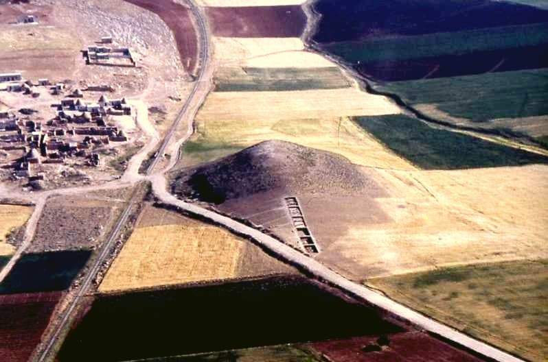 Tell Karamel'in havadan görünüşü: ekili tarlaların ortasında höyük ve yamacındaki kazı açması, solda köy, köyle höyük arasından geçen demiryolu ve yol