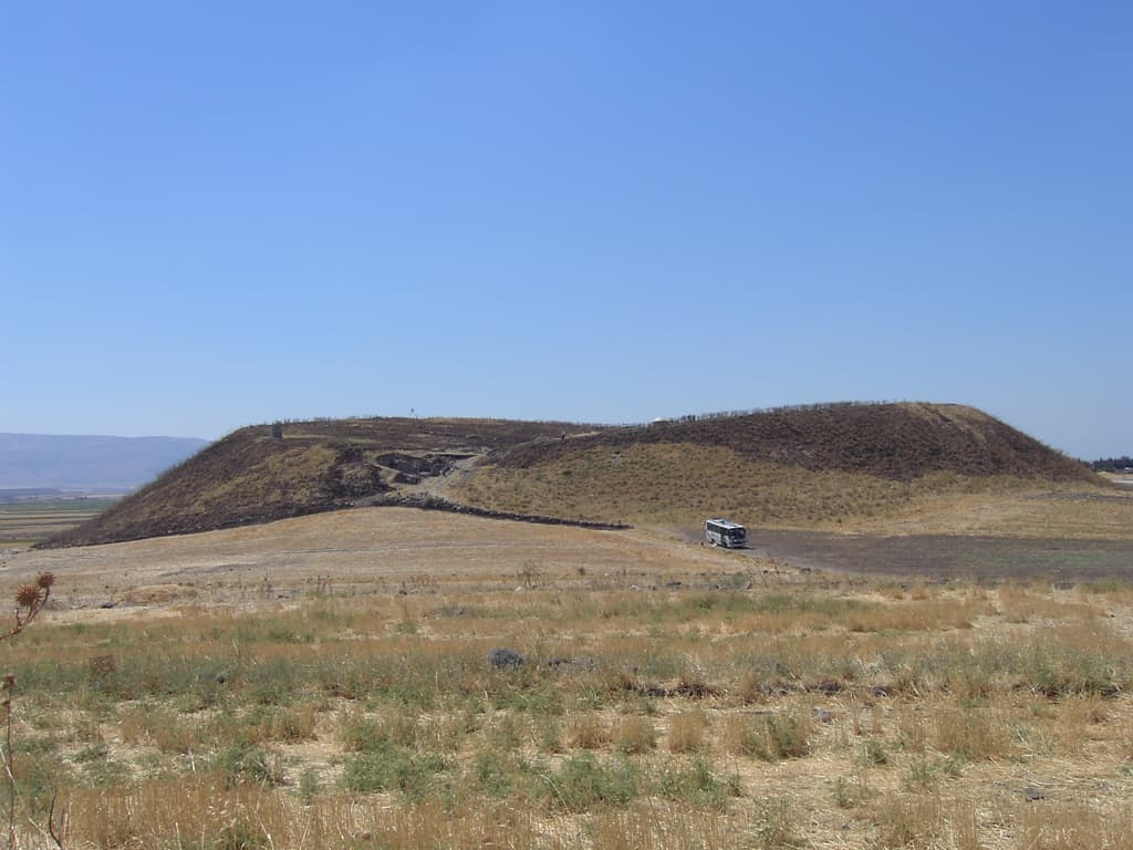 Tell Karkur'un yüksek höyüğü, kuzeydeki alçak höyükten güneye bakış; kuru otlu ovadan yükselen koyu renkli höyük ve önünde küçük bir otobüs